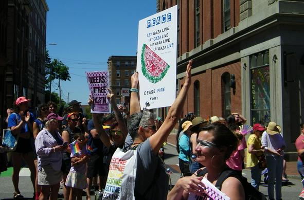 Someone holding up a sign at a Pride parade that reads:
PEACE
LET GAZA LIVE
LET GAZA LIVE
LET GAZA LIVE
LET GAZA LIVE
CEASE FIRE NOW!
On the sign is an image of a green and red watermelon slice. 
The person is wearing a white cape with the words PALESTINE written in all the colors of the rainbow.
Another person holds up a sign that reads: Queers for a Free Palestine.