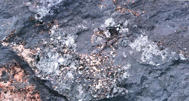 Overall view of shiny metallic masses, with plate like crystals in areas. Dark rock surrounds them.