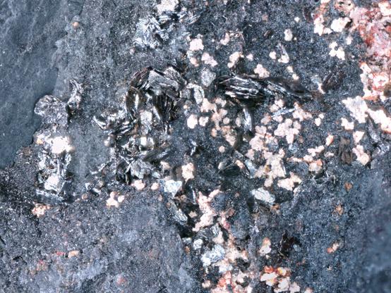 Close up view of hexagonal silver crystals surrounded by dark matrix rock and pink clay and oxides.