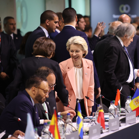 President von der Leyen shakes hands with a participant to the Summit. They are both about to sit down in a room full of people. In front of them there are flags of the represented countries.