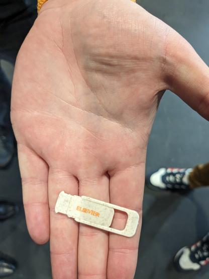 Foto einer offenen Handfläche, auf der so eine anklebbare, auf und zu schiebbare Abdeckung für in Laptop verbaute Kameras liegt. Die Abdeckung ist mit "Elsevier" gebrandet.