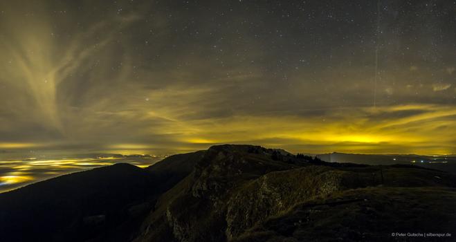 Ein dunkler Bergrücken bei Nacht, hinter dem links und rechts Teile der weiter entfernten Landschaft zu sehen sind, Links ein Nebelmeer, unter dem gelbliche Stadtlichter hervorleuchten. Am Himmel sind Sterne zu sehen, aber auch großflächige Zirruswolken, die das Licht der Städte reflektieren.