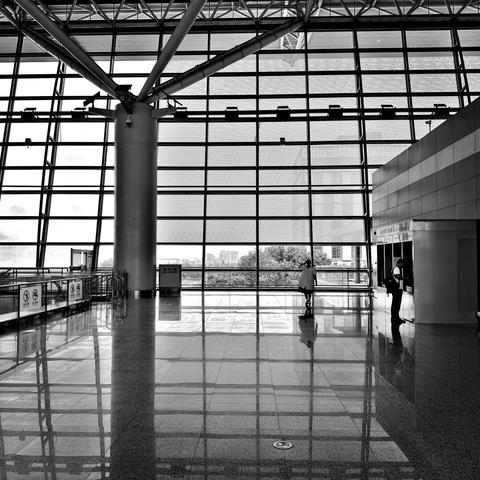 An airport’s multi-panelled window in the distance, with reflections from it and a large pole on the ground in front. The photo is black and white. A person stands at an ATM in the distance over to the right, and someone looks out of the window towards the middle.