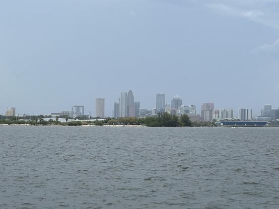 A daytime view of the Tampa skyline, taken from a ferry across the water. The foreground features the calm waters of Tampa Bay, with a strip of land covered in greenery and small buildings along the shoreline. The background showcases the tall buildings of downtown Tampa against a clear, slightly overcast sky. The mix of modern skyscrapers and older structures creates a dynamic cityscape, while the overall scene conveys a sense of tranquility and urban beauty.