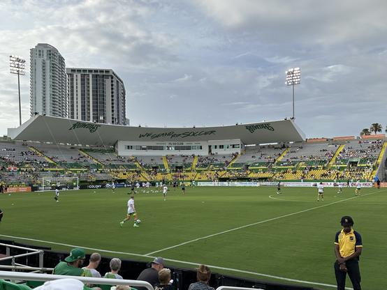 A lively scene at a soccer stadium during the day, featuring the Tampa Bay Rowdies' home ground, Al Lang Stadium, in St. Petersburg, Florida. The stadium is filled with spectators, and players are seen on the field warming up. The stands are decorated in green and yellow, with some fans wearing matching team colors. The words 'WE STAND FOR SOCCER' and 'ROWDIES' are prominently displayed on the stadium's structure. In the background, tall buildings rise against a partly cloudy sky. A security guard stands near the field's edge, overseeing the event.