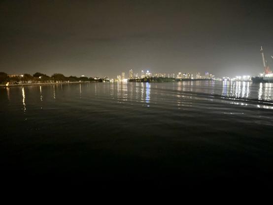 A nighttime view from a ferry on the water, looking towards the city skyline of Tampa, Florida. The city's lights are reflected in the calm, dark water, creating a shimmering effect. On the left side, there is a line of dimly lit trees and buildings along the shore. On the right, some industrial structures are faintly visible under bright lights. The sky is clear and dark, adding to the serene atmosphere.