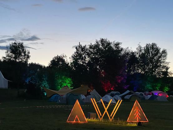 Foto des neuen dreiteiligen Aufstellers des VVoidcamp Logos, dahinter bunt beleuchteter Wald und langsam dämmrig werdender Abendhimmel