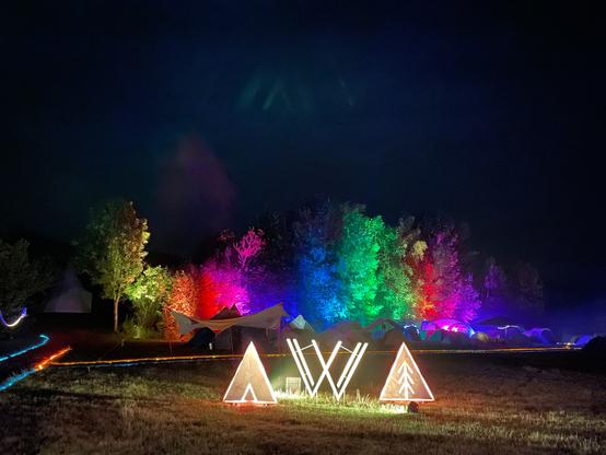 Foto des VVoidcamp Logo Aufstellers bei Nacht. Er leuchtet hell warmweiß vor dem bunt beleuchteten Waldstück