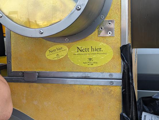 2 Sticker an einer Wand. Der größere ist ein "Nett hier. Aber waren sie schon mal in Baden-Württemberg?" Sticker. Daneben der kleinere "Nett hier. Aber waren sie schon mal auf den MRMCD?" Sticker.