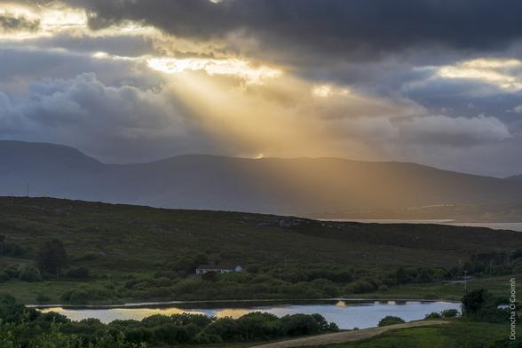Light pours from a gap in the clouds near sunset over Sheep's Head. There's a small lake in the foreground, reflecting the light, and a bungalow sits near the lake.