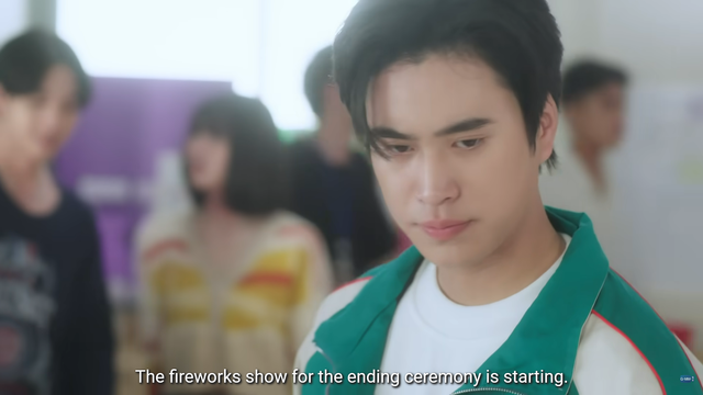 Gemini as Krongtap stands in the foreground of a classroom. Other people are in the background, but are very blurry. 

Of note, there is a window in the background. It's also blurry and indistinct, but it looks to be nearly white with sunlight shining in.

There's a caption as someone of screen that says: "The fireworks show for the ending ceremony is starting."