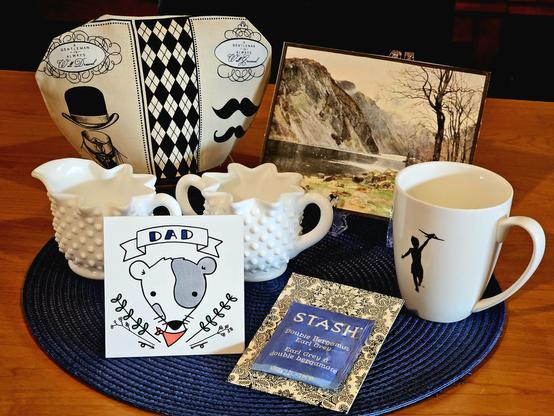 A tea tableau in sedate colors of beige, blue, and black with a Father's Day theme. There is a drawing of a dog with a "Dad" banner, a mug with an image of a boy in silhouette flying a toy airplane, a tea cozy in a bowler hat & mustachio print fabric, a watercolor of a mountain stream, and Double Bergamot Earl Grey tea from Stash Teas.