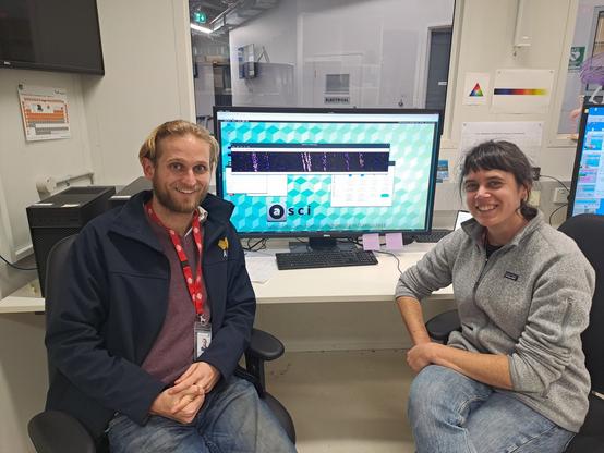 2 people - a man and a woman - sitting side by side and smiling at the camera. Between them is a computer monitor showing some tantalising data - bright lines against a dark background. You'll have to wait and see to find out what it all means!