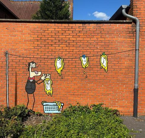 Streetartwall. A funny comic-style mural has been sprayed onto a long red stone wall. It is a simply drawn little man with a protruding lower jaw, one large and one small eye, thin arms and legs in a black suit with a red bow tie. He stands cheerfully against the wall with his mouth open and hangs melted, deformed clocks on a washing line. He takes the clocks from a laundry basket. It is no coincidence that the clocks are reminiscent of a Dali painting. The artist has sprayed 7 walls in homage to great artists.