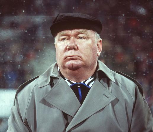 De legendäre ukrainische Fußballtrainer Walerij Lobanowskyj auf einer Trainerbank sitzend. Mit schwarzem Käppi und grau-braunem Mantel verfolgt er scheinbar teilnahmslos ein Spiel seiner Mannschaft.