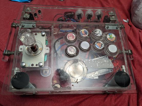 overhead view of an arcade style fight stick controller. The 8 buttons are arranged in an ergonomic shape. The entire body of the arcade stick is made from clear plastic and red fabric is visible beneath it. The joystick is also clear

Some of the buttons feature pictures of fighting game characters

top row: Marisa (Street Fighter 6), Birdie (Street Fighter 5), Hakumen (Blazblue), Piccolo (DragonBall FighterZ)

bottom row: Mila (Dead or Alive 6), none, Dudley (Street Fighter 4), none