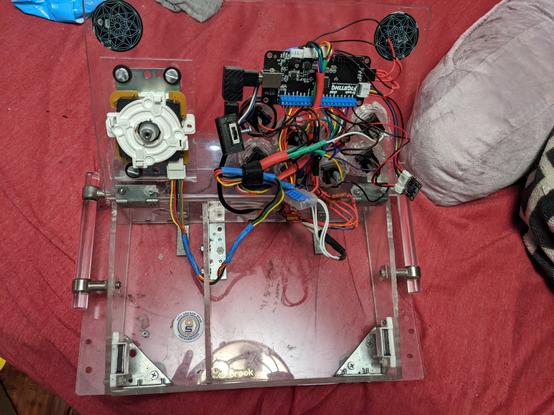 overhead view of an arcade style fight stick controller. The entire body of the arcade stick is made from clear plastic and red fabric is visible beneath it. The top panel has been flipped up to reveal that all Yue wiring and electronics are attached to that top panel