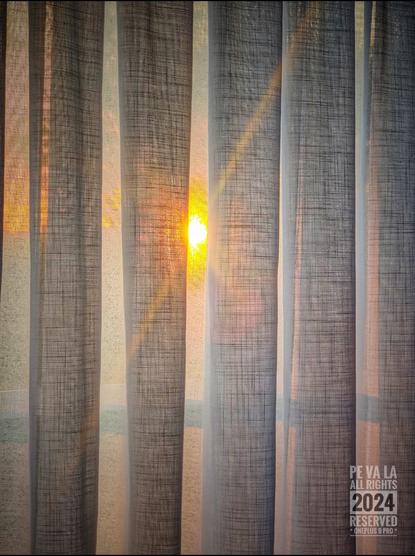 Kleurenfoto genomen van een zonsondergang doorheen een standaard (wit) gordijn.
Door de houding bij het nemen van de foto is er een lichte flare van de ondergaande zon.
Door de zonsondergang is de lucht ook mooi rood.