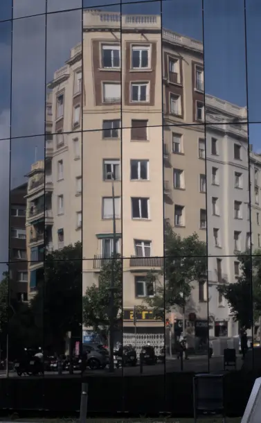 A photograph of the reflection of a building in the glass panes of another building.  The glass building has a curved surface made up of tall, straight, flat glass planes linked together at an angle to form the curve.  This has the visual effect of 'breaking up' any large reflection that would take multiple panels; some panels have a brighter reflection, some are darker or almost cloudy, and due to the angle difference some pieces of the reflected building are jutting up uncomfortably against sky or different parts of the building they don't encounter in real life; it's as if they took the 8 story slate brown building with dark brown walls on the top two floors and cut out various close-but-not-contiguous columnar regions, then re-arranged them into a somewhat flat surface.  Note that the distortion/cutout effect is _only_ vertical; any given column of glass panes has the same angle, so the reflection going up from any point is continuous.  The building being reflected, in reality, is at an angle here (it's a building on a corner, so it has at least two faces exposed to the street), but it's hard to tell.  Various sections of trees are visible in the panels as well; some sections are brighter and others are dark.  They have the same cut at random effect as the building itself.