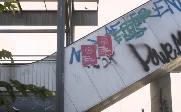 A photograph of the outer enclosed part of a pedestrian overpass stairwell; on the left is some metal fencing and fence roofing, with part of a tree visible, and a tiny bird on the root.  On the right is the enclosed stairwell, looking like a tube rising to the upper right of the image, and part of a support column and fence visible below it.  The sky in the background is bright with a golden hue.  Dirt is visible and stains the enclosure.  A minor tagging war has been going on, with some tags painted over with other ones.  Someone who goes by the tag of "nob" has written their name twice: once on the grey metal of the support on the right, and once on the brighter, dirty cream colored enclosure.  One of the tags is in black and large letters (another is black but small).  The remaining tags are blue and green.  Rising with the enclosure, not too far from the top, are two red posters for a political party.  They have a logo that looks like a four pointed, dual shaded star (like an origami star) with a circle behind it, slightly eclipsed by the star; surrounding that is a ring of 12 white five pointed stars.  In bold text underneath this is the phrase CONTRA LA GUERRA I L'AUSTERITAT (AGAINST WAR AND AUSTERITY), with the letters OJS at the bottom, likely an acronym for the party itself.  Smaller text can be seen but not read in various places in the poster.
