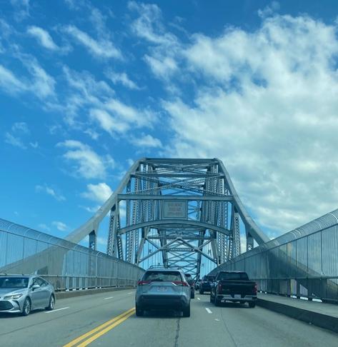 View from my car as we approach the Bourne Bridge in Massachusetts. It is a steel arch bridge with a suspended roadway. Overhead, puffy white clouds dot the blue sky.
