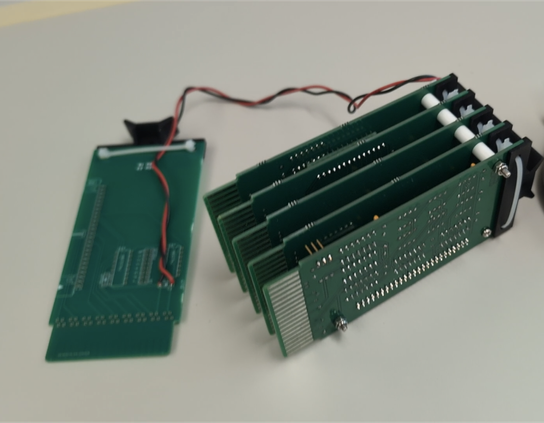 Five green printed circuit boards about the size of a 3x5 card are bolted together in parallel like a five-layer cake. A sixth card has a pair of wires running from it to the five-card stack.