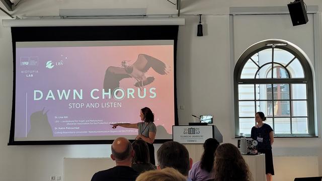 On the right are two women, the left one is pointing at the screen. On the screen is a colorful slide with a flying bird and the words "dawn chorus"