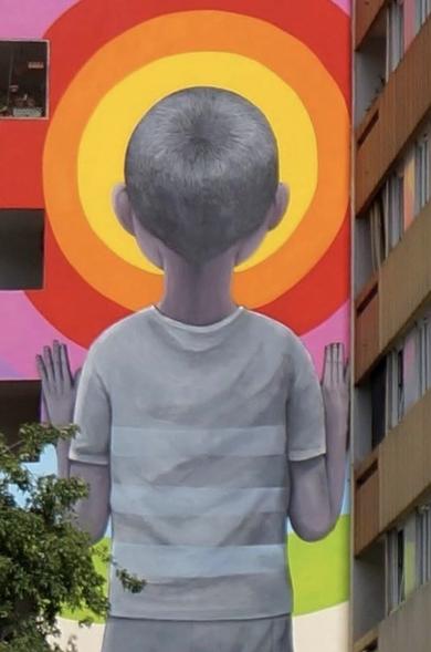 Peinture murale d'un enfant aux cheveux courts et au t-shirt rayé en nuances de gris, face à un fond formé de cercles aux multiples couleurs vives sur le côté d'un immeuble.
