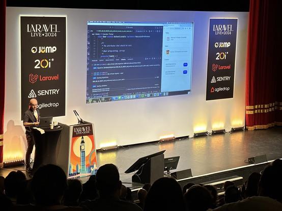 Dan Johnson presenting at Laravel Live 2024, with a code editor and messages displayed on a large screen behind them. The stage features banners with logos, and the audience is visible in the foreground.