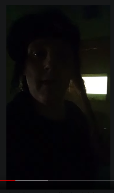 Screenshot of Pandora from her second video. She is in a darkened cell, with diffused daylight from a frosted window to outside the only light in the room, looking at the camera and in clearly visible emotional distress. It is too dark to see much detail of her face, but she is wearing a dark coloured hat and dark t-shirt, and on her face is an expression of sadness and distress.