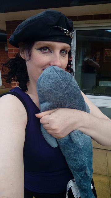 Pandora taking a selfie. She is standing in a courtyard, in front of a brick wall and a large clear window. She is wearing the same hat from her first video, and a dark blue top. She has shoulder-length curly black hair, is wearing dark eye make up, and is hugging a Blahaj and smiling.