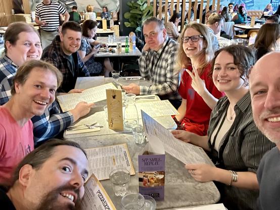Group of seven people sitting around a table at a restaurant, smiling and holding menus.