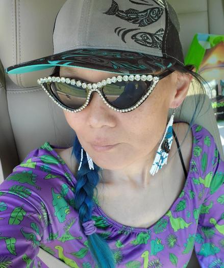 Selfie in car. Wearing my cateye rhinestones sunglasses, Indigenous wild salmon art cap hat & beaded raven earrings & purple/green dinosaurs & plants top. Blue hair is braided & hanging over my right shoulder.