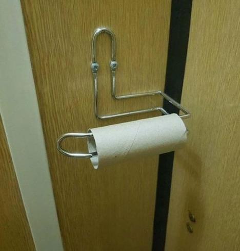 A metal toilet paper holder in a corner od a bathro,with an empty roll, that looks similar to a large paperclip