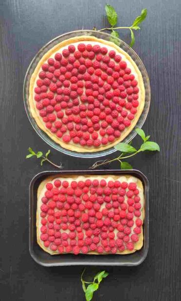 Deux tartes aux framboises sur un table noire, la première est dans un plat rond, la seconde dans un plat rectangulaire. Des brins de menthe épars entre et autour des plats.