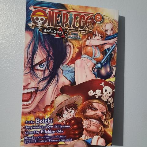 Photograph of a white person's hand holding Volume 2 of One Piece Ace's Story The Manga. The cover shows half of Ace's face on the left side with various One Piece characters drawn throughout the rest of the cover (Nami, Kalifa, Roger, Luffy). Credits are in the lower left hand corner listing the artist as Boichi and the storyboard by Ryo Ishiyama.