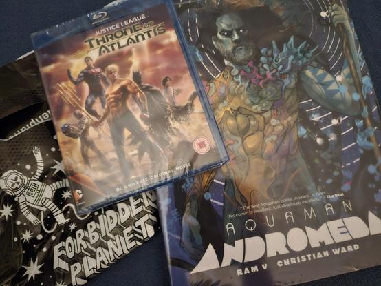 Die Bluray zu "Justice League: Throne of Atlantis" und das Buch zu "Aquaman: Andromeda" liegen auf einer blauen Tischdecke. Halb verdeckt: eine Einkaufstüte der Comicladen-Kette "Forbidden Planet".