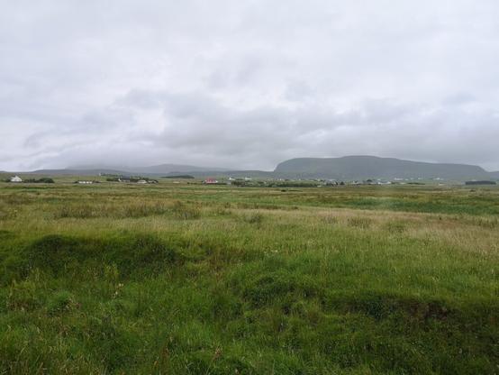 Wir sehen über eine weite flasche Landschaft in Schottland auf der Isle of Skye. überall sieht man grünes Gras und in der landschaft verteilt weiße Häuser. Ganz in der Ferne sieht man ein Bergmassive, auf das weiße Wolken sitzen. Der Himmel ist bewölkt und ehr grau