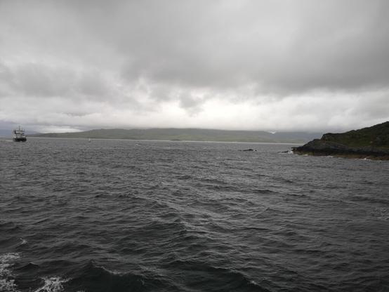 Wir sehen ein graues Schwarzweiß Bild vor uns. Die Sicht geht auf den Atlantik hinaus, wo man rechts noch einen Felsen im Meer sieht und von link kommt eine Fähre auf uns zu gefahren. Das Wetter war an dem Tag sehr feucht und nass und sehr trüb