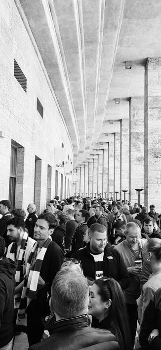 Black and white photo of a concourse at the Olympic Stadium in Berlin,  Germany during the half time break. Fans are queuing for beers and other refreshments. Some are already enjoying theirs. The concourse swerves to the left with pillars.