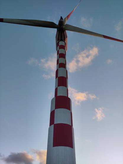 View looking up at a large wind turbine, painted in red and white checks against a blue sky with small grey and pink clouds at sunset.