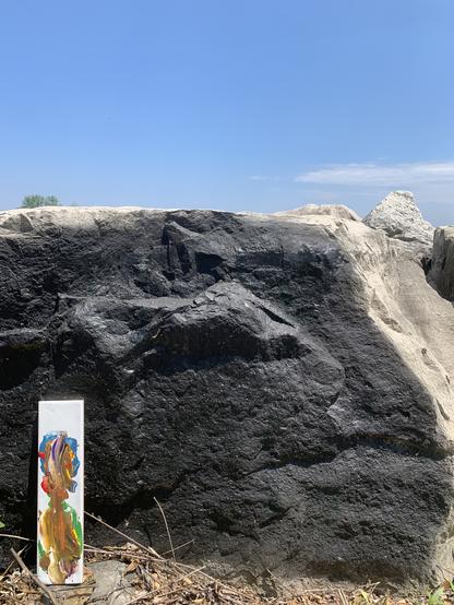 A rock painted black has a white rectangular tile with splashes of coloured paint in front of it. In the background above, a blue sky with wispy clouds