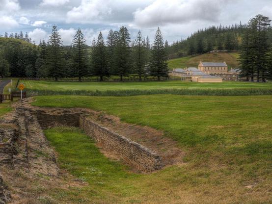 In the foreground, the rectangular stone-lined sawpit where convicts cut the timber, across the grassy field the old stone military barracks. Also, a whole lotta Norfolk pines.