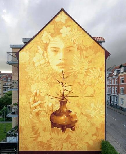 Streetartwall. On the outside wall of an old two-storey building a beautiful mural of a young woman and a tree was sprayed/painted. Reduced to the warm colors yellow and brown, the face of a young woman is depicted disappearing pale in a pale yellow foliage and bright flowers. She appears almost fragile as she raises her hand protectively over a thorny little tree in a golden-brown clay pot. The clay pot is typical of Chilean art from the Atacama Desert and has a small cow's head on the front. The tree (a Prosopis tamarugo tree), also stands for Chile and the unconventional beauty of a plant that thrives even in the driest regions. A symbol of life and resistance for the local population. Extraordinary.