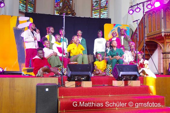 Eine junge Frau und ein junger Mann singen, die Kinder sind um sie herum platziert.