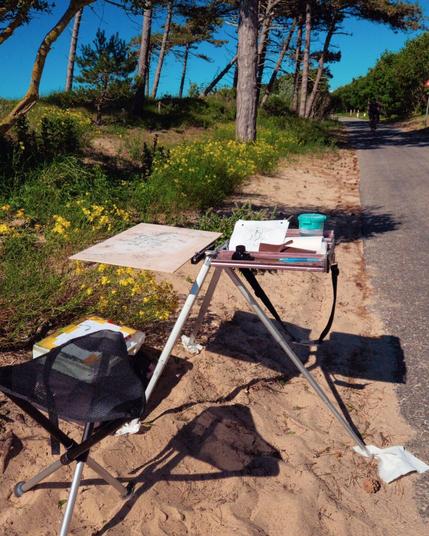 Easel standing in the sand next to a pine tree.