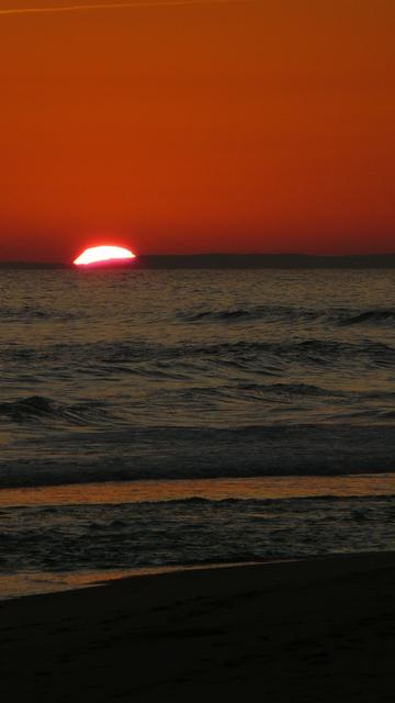 A bright white sliver of the setting sun over the dark blue waters of the Atlantic ocean against  red skies at Porto de Mós beach in the summer solstice of 2011