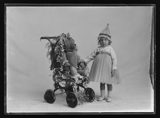 Black-and-white photo: Daffodil Day Fancy Dress Entrant. 16 September 1916. Photographer, F. N. Jones. Description: Full length studio portrait of a young girl in fancy dress with a teddy bear seated in a decorated push chair. Citation: The Nelson Provincial Museum, Photo Collection Reference No. 311530. https://collection.nelsonmuseum.co.nz/objects/P25951/daffodil-day-1691916-fancy-dress-entrant