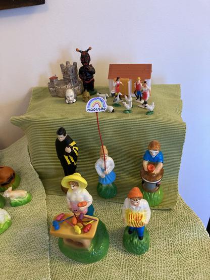 A "cascata" display of colorful hand-painted figurines on a landscape of green and blue cloth. In the center one figure holds a rainbow sign reading ORGULHO (Pride).