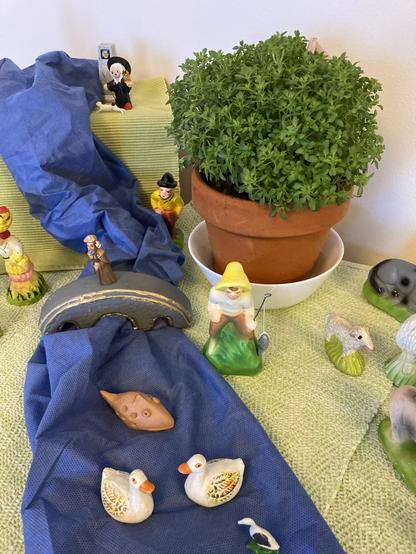 A "cascata" display of colorful hand-painted figurines on a landscape of green and blue cloth. A small round St. John's basil plant is in the middle.